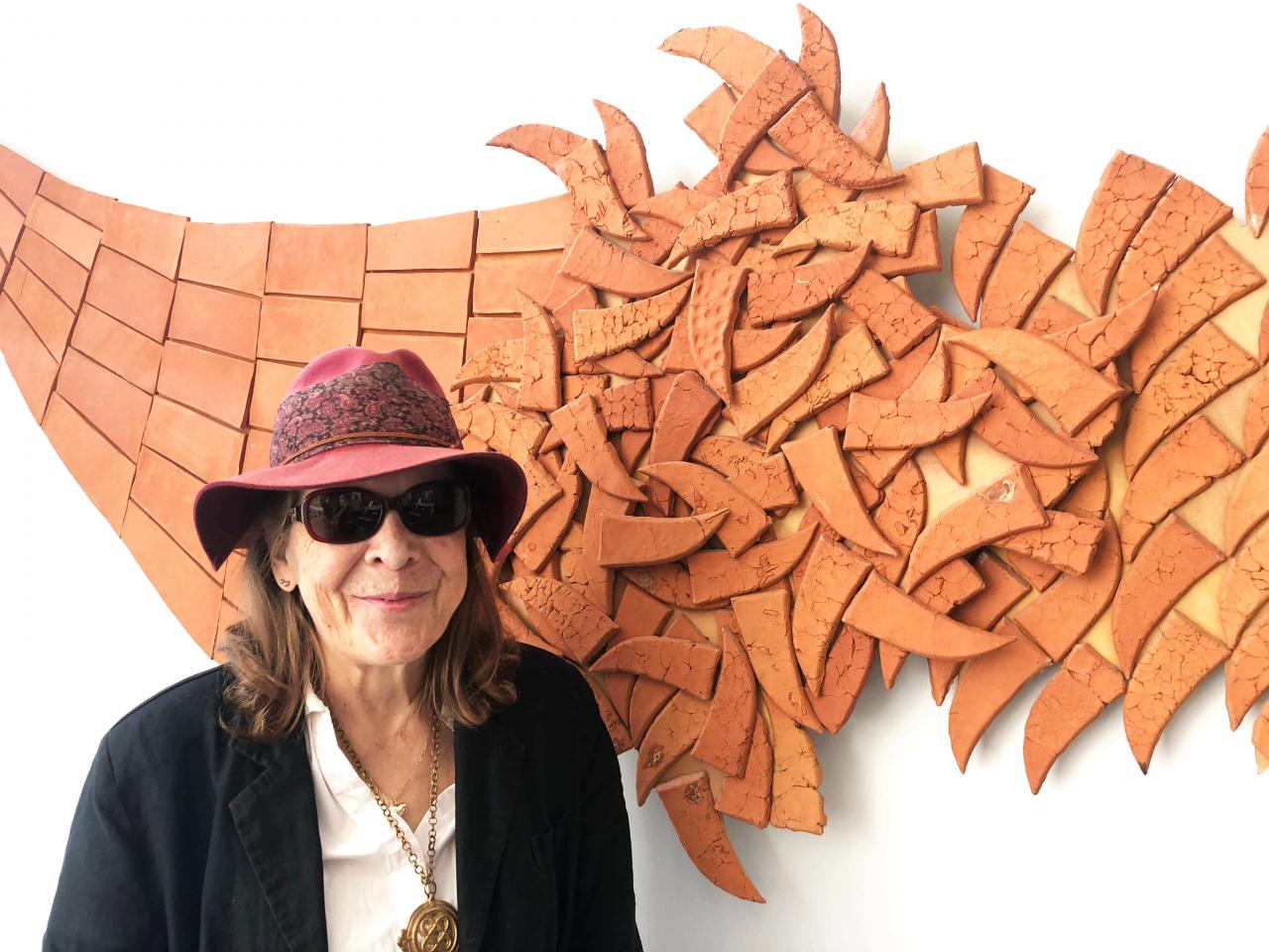 Ruskeista savitiilistä koostuva taideteos, ja edessä aurinkolaseissa ja huopahatussaan hymyilevä henkilö, jonka ruskeat hiukset ulottuvat olkapäille.